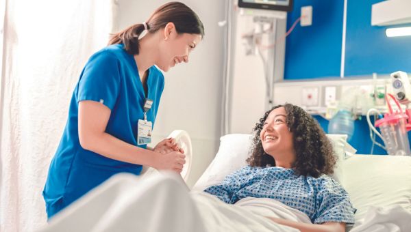 A nurse wearing blue scrubs with her hair in a low ponytail attending to smiling hospital patient in hospital bed.