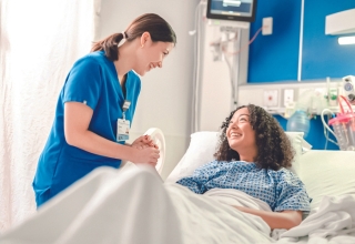 A nurse wearing blue scrubs with her hair in a low ponytail attending to smiling hospital patient in hospital bed.