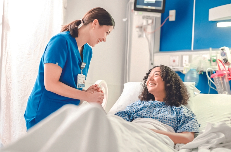 A nurse wearing blue scrubs with her hair in a low ponytail attending to smiling hospital patient in hospital bed. 