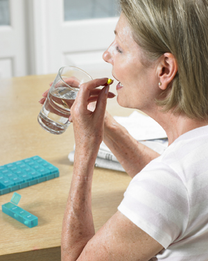 Woman sitting at table taking pill, holding glass of water. Pill organizer is on table.