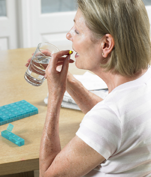 Woman sitting at table taking pill, holding glass of water. Pill organizer is on table.