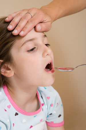 Girl being given medicine in spoon, adult hand on her forehead.