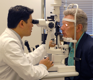 Man sitting in front of eye exam scope. Man's chin is resting on support. Healthcare provider is sitting on other side of scope, looking through eyepieces to examine man's eyes.