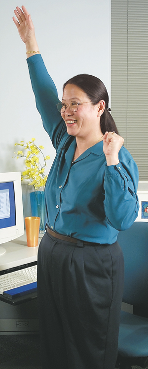 Person standing at workplace desk, back straight, stretching one arm straight above head.