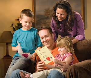 Family, children and wife gathered around Dad, reading handmade greeting card to Dad.