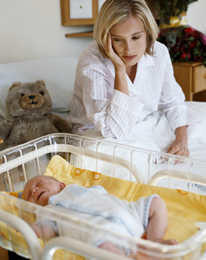 Woman sitting on bed looking at newborn in crib.