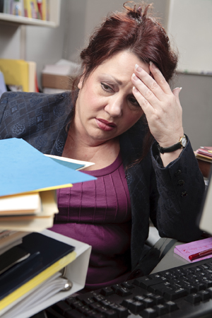 Overweight woman in an office cubicle behind a stack of folders and papers, holding head, under stress.