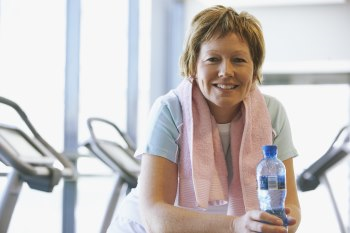 Photo of a middle aged woman at the gym. She is looking at the camera and holding a water bottle. Exercise machines are lined up behind her.