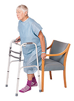 Image of woman with a walker backing up into chair - one hand on walker and one on the arm of the chair - the chair is touching the back of her legs.