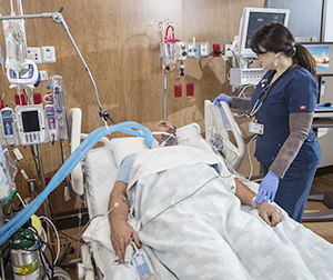 Patient lying in intensive care unit bed with medical machines surrounding him. Tube in his mouth from ventilator. Health care provider is standing by the bed.