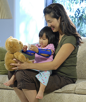 Toddler with arm restraints playing with teddy bear while sitting in woman’s lap.
