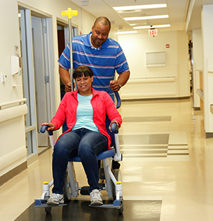 Man pushing woman in wheelchair in hospital hallway.