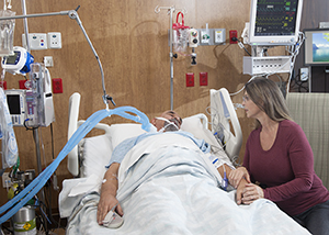 Woman sititng next to bed of intubated man in intensive care unit.