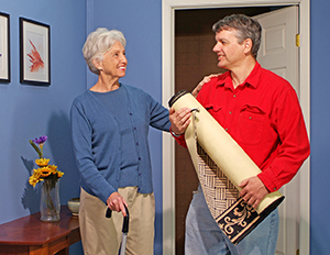 Man holding rolled-up area rug, talking to woman with cane.