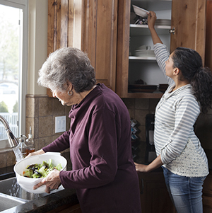 Woman reaching into upper shelf in kitchen cabinet while older woman makes salad.