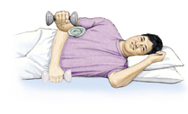 Man lying on side with head supported by pillows. Hand is grasping hand weight with elbow bent and weight against stomach. Rolled-up towel is under elbow. Ghosting shows man lifting hand weight up with elbow remaining in place.