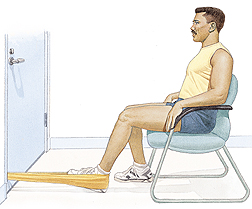 Man sitting in chair facing doorway. Plastic tubing is looped around heel of one foot, ends of tubing shut in door. Man is pulling leg back to stretch tubing.
