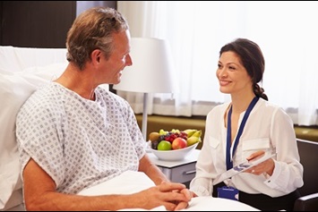 A female healthcare worker is seated while talking to a male patient who is in a hospital bed.