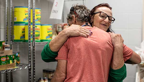 two women hugging next to shelves of food at food pantry