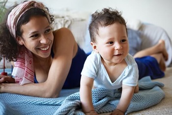 Childhood, physical development and childcare concept. Picture of attractive cheerful young mixed race female laughing, watching her adorable toddler child making first crawling movements on floor