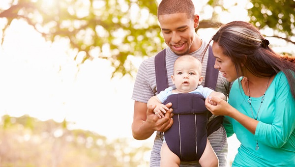 Family With Baby Son In Carrier Walking Through Park