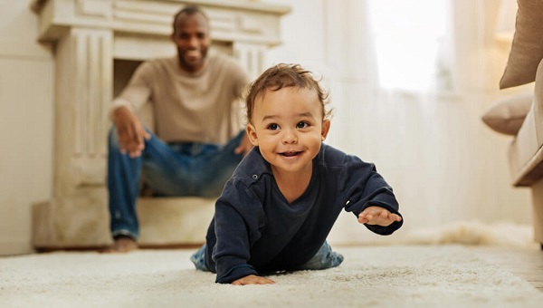 Happy childhood Happy darkhaired afroamerican man laughing and watching his cheerful young son crawl