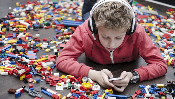 A boy is looking at his cellphone and listening to his headphones while surrounded by Lego bricks on the floor.