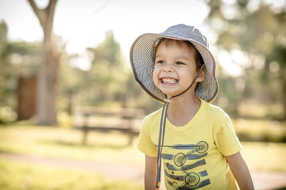 A smiling boy is wearing a large hat outdoors to protect his face from the sun