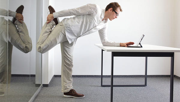 A man is standing and stretching one leg while looking at his laptop at work