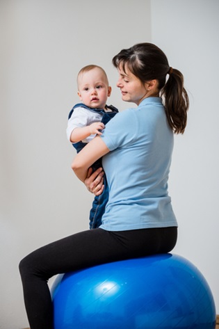 A mom sits on a large exercise ball while holding her baby.