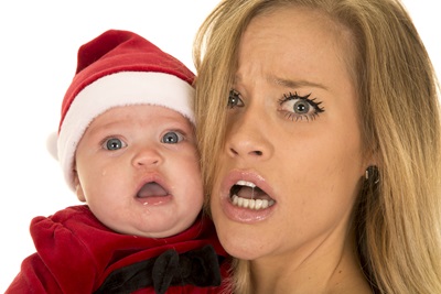 A mom holds her baby, who is wearing a Santa Claus outfit. Both have shocked expressions on their faces.