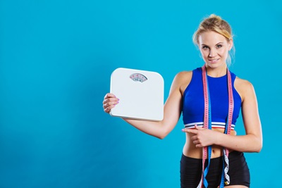 A woman is wearing exercise clothes and holding a weighing scale and tape measures.