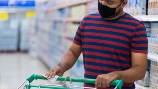 Asian man wearing medical face mask wipes shopping cart handle in supermarket