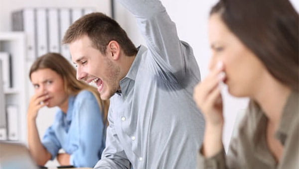 During a work meeting, a young man raises his left arm and reveals a sweat stain in the armpit area of his shirt. Two of his female coworkers hold their noses.