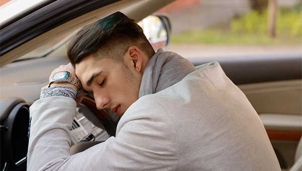A young man is napping in his car and resting his head on the steering wheel.