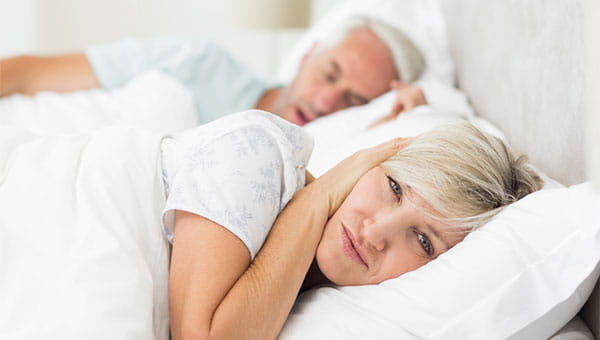 A couple in bed with one person snoring and the other with their hand over their ear.