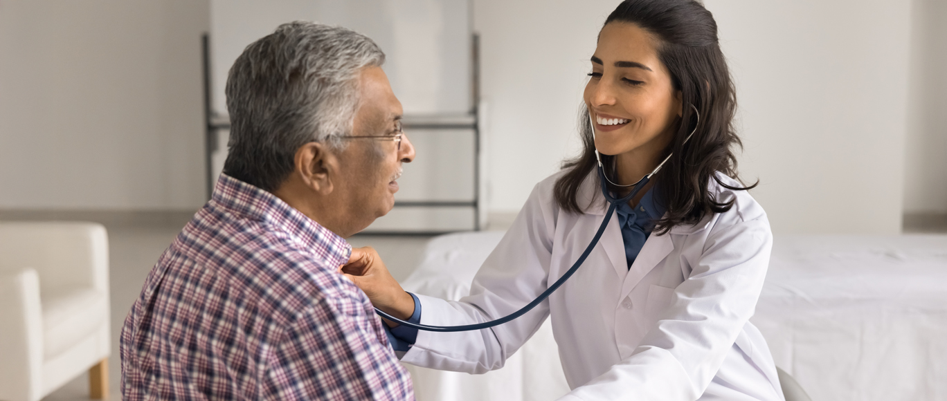 A female primary care doctor listens to the heart of a middle-aged man through a stethoscope.