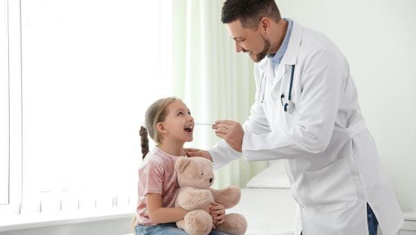 A young girl holds a teddy bear while a doctor takes a swab of the inside of her mouth.