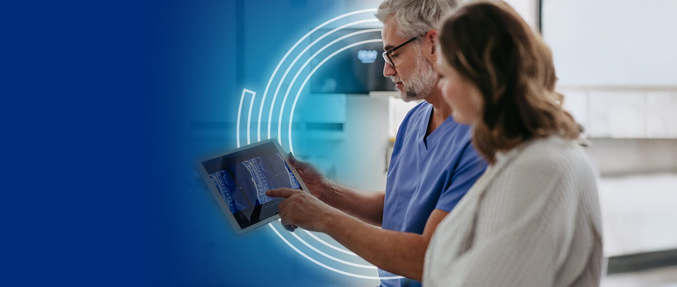 A doctor in scrubs shows a patient x-ray on a tablet to another doctor in a lab coat.