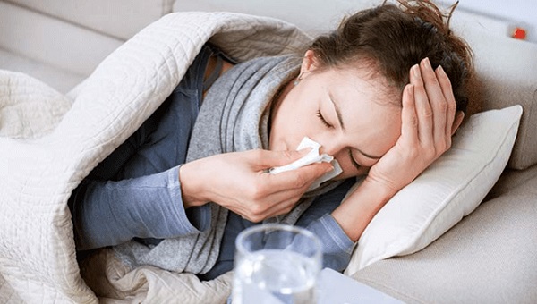 woman holding her nose with a tissue laying in bed with hand on forehead
