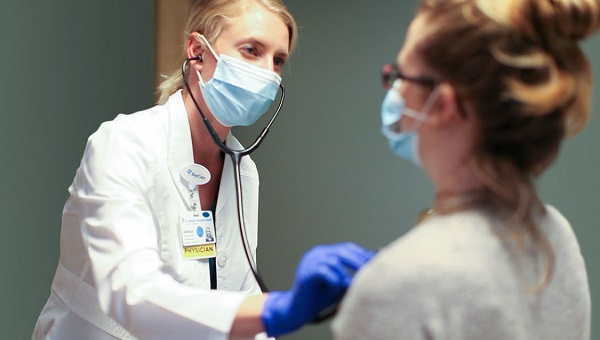 A female physician, who is wearing a mask, is examining a female patient who is also wearing a mask.