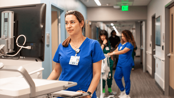 A BayCare nurse in blue scrubs stands by a monitor in a medical hallway.