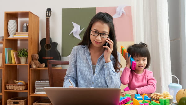 Asian woman with smartphone using laptop while little girl having fun with colorful blocks pretending