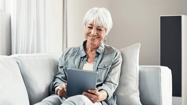 A senior woman is sitting on her sofa while looking at information on her tablet.