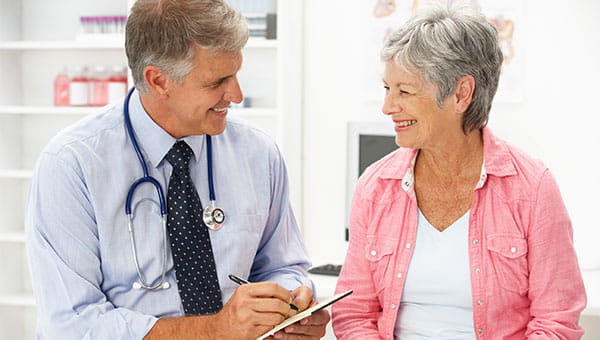 Male doctor with a notepad and pen smiling and sitting next to an older female patient with short grey hair smiling at him