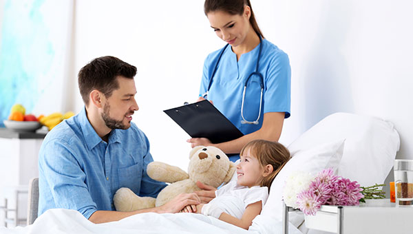 A girl patient getting a teddy bear from her father while a female nurse stands nearby