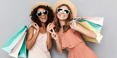 Two young women smiling, wearing sunglasses and holding shopping bags over their shoulders