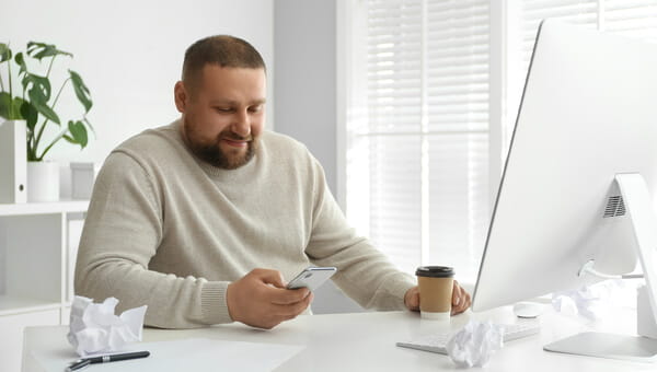 Man looking at a phone in front of a computer with a cup of coffee