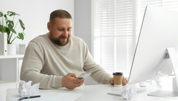 Man looking at a phone in front of a computer with a cup of coffee