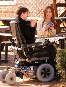 Two people having coffee outside at a picnic table, one of them in a motored wheel chair